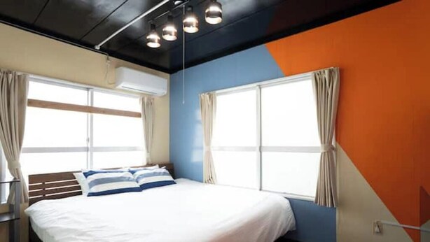 A hotel room with a bed, two windows with curtains, a ceiling light fixture, and a partial view of a wall with a geometric design.