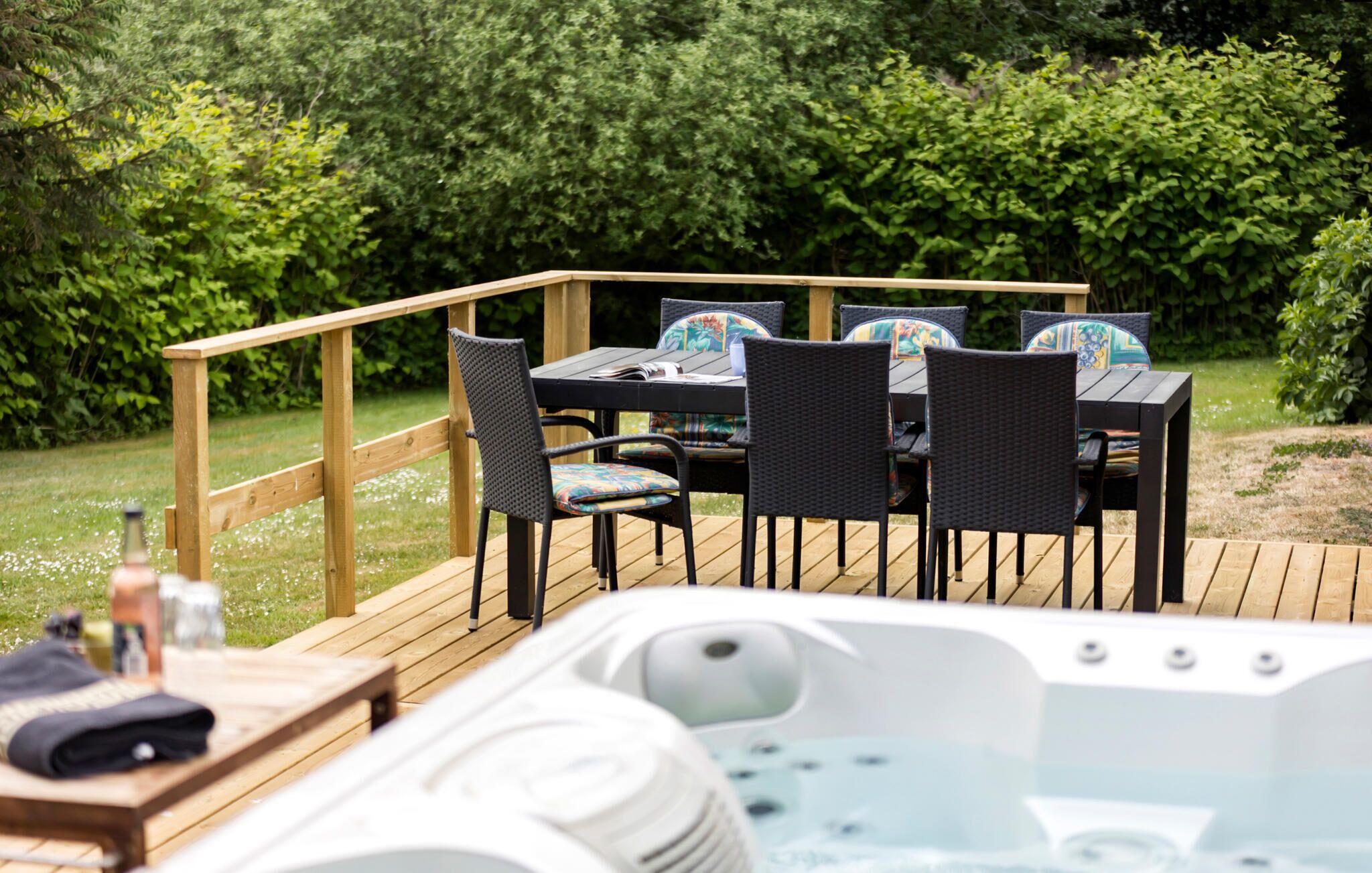 Eine Holzterrasse mit Whirlpool, Gartenmöbel mit Korbstühlen und Kissen sowie einem Holztisch.