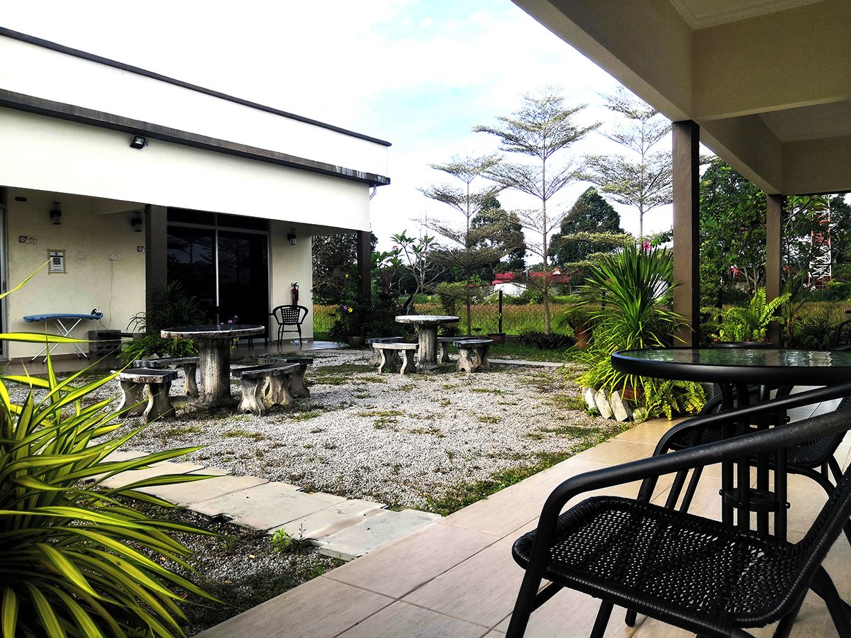 An outdoor patio area with stone tables and chairs, surrounded by greenery and a building with a white facade.