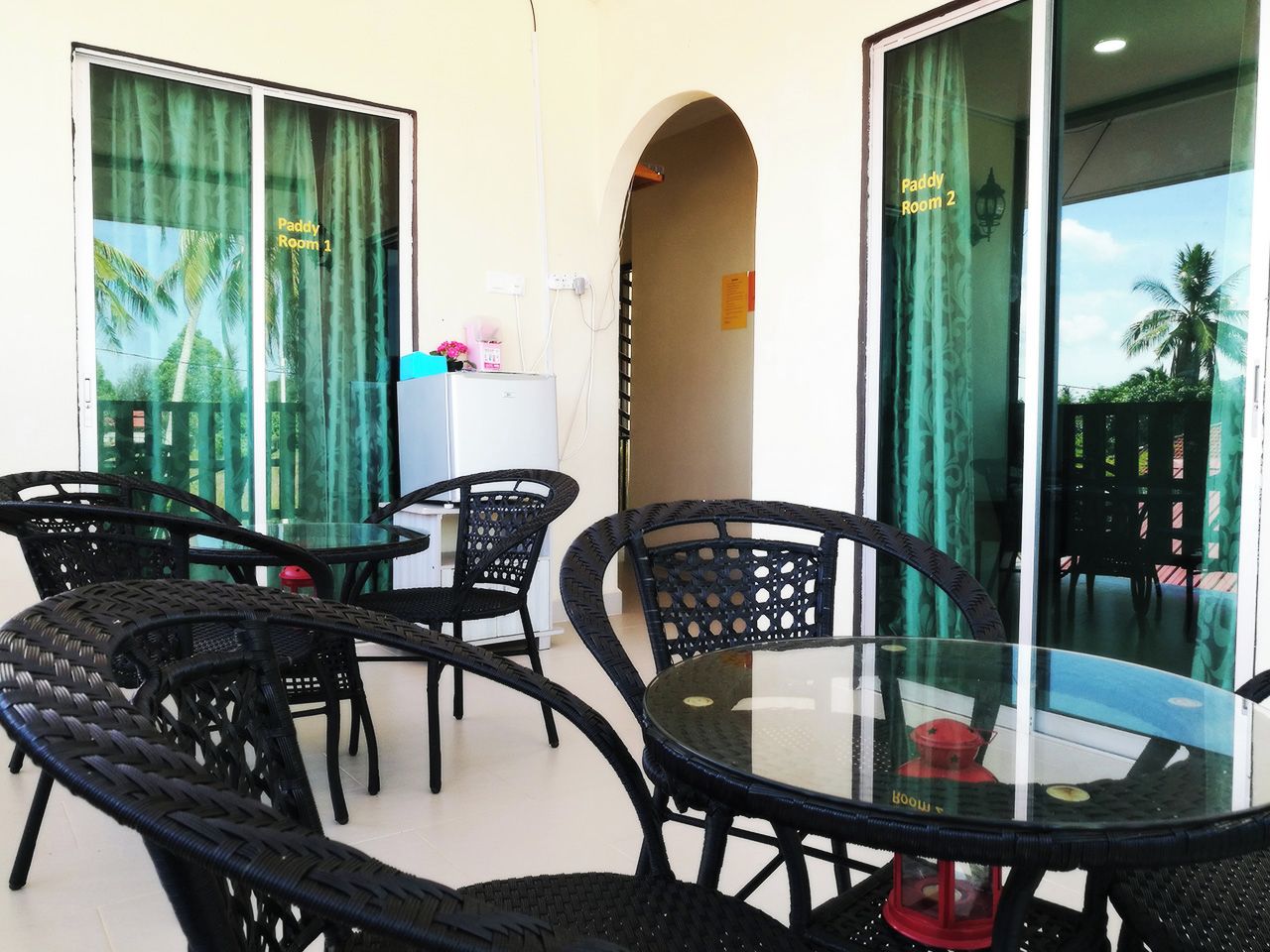 Une pièce avec une table et des chaises en verre, un vase rouge et une vue sur des palmiers à travers les fenêtres.