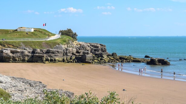 A coastal landscape with a sandy beach, rocky cliffs, and a grassy hill with a building and flag. - Bel Appartement Proche Mer (Royan)