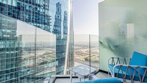 Um edifício alto com fachada de vidro, uma varanda com sofá azul e uma pequena mesa, e vista para a paisagem urbana.