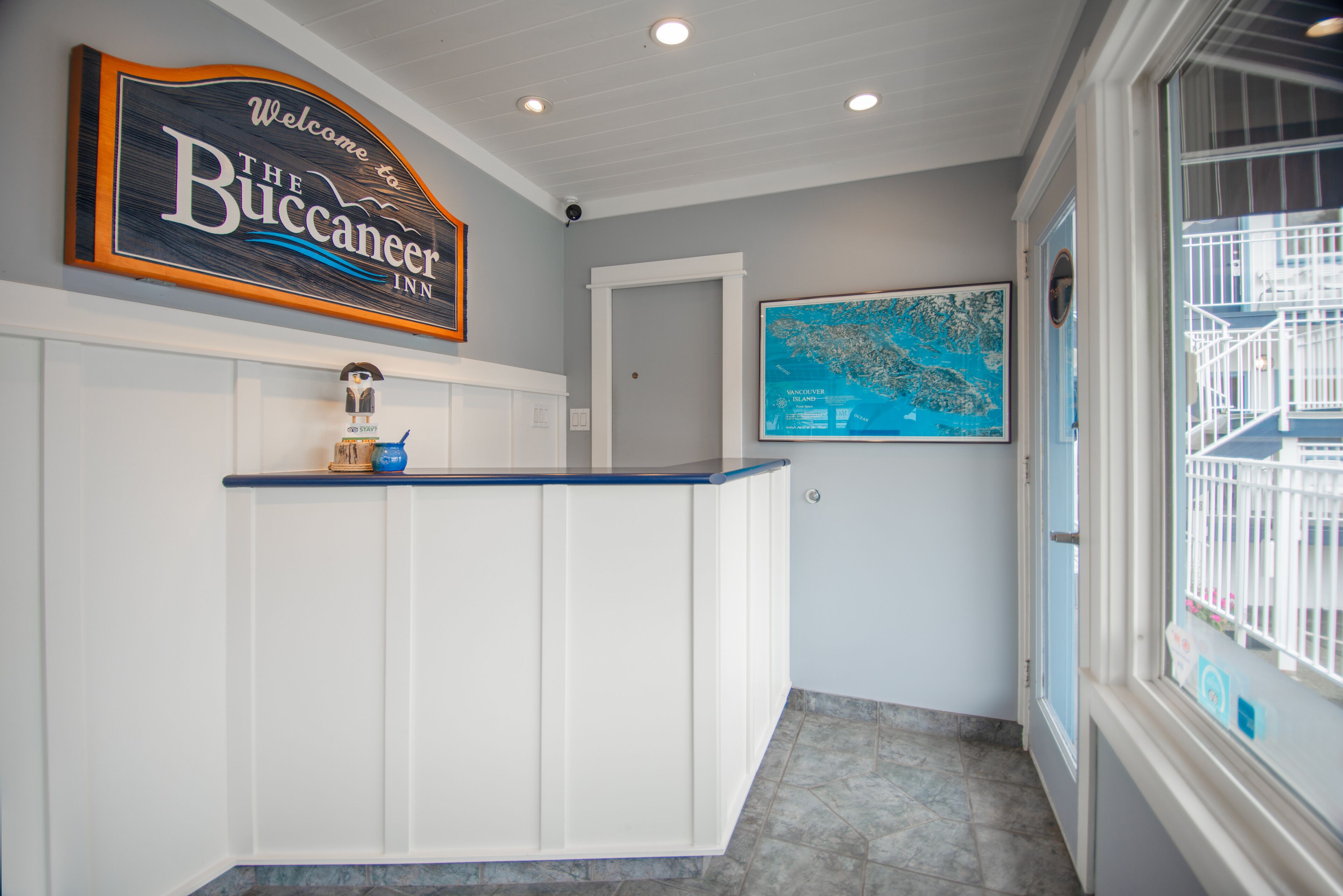 A reception area with a sign reading "Welcome to The Buccaneer Inn," a reception desk, a framed map on the wall, and a door leading to another room.