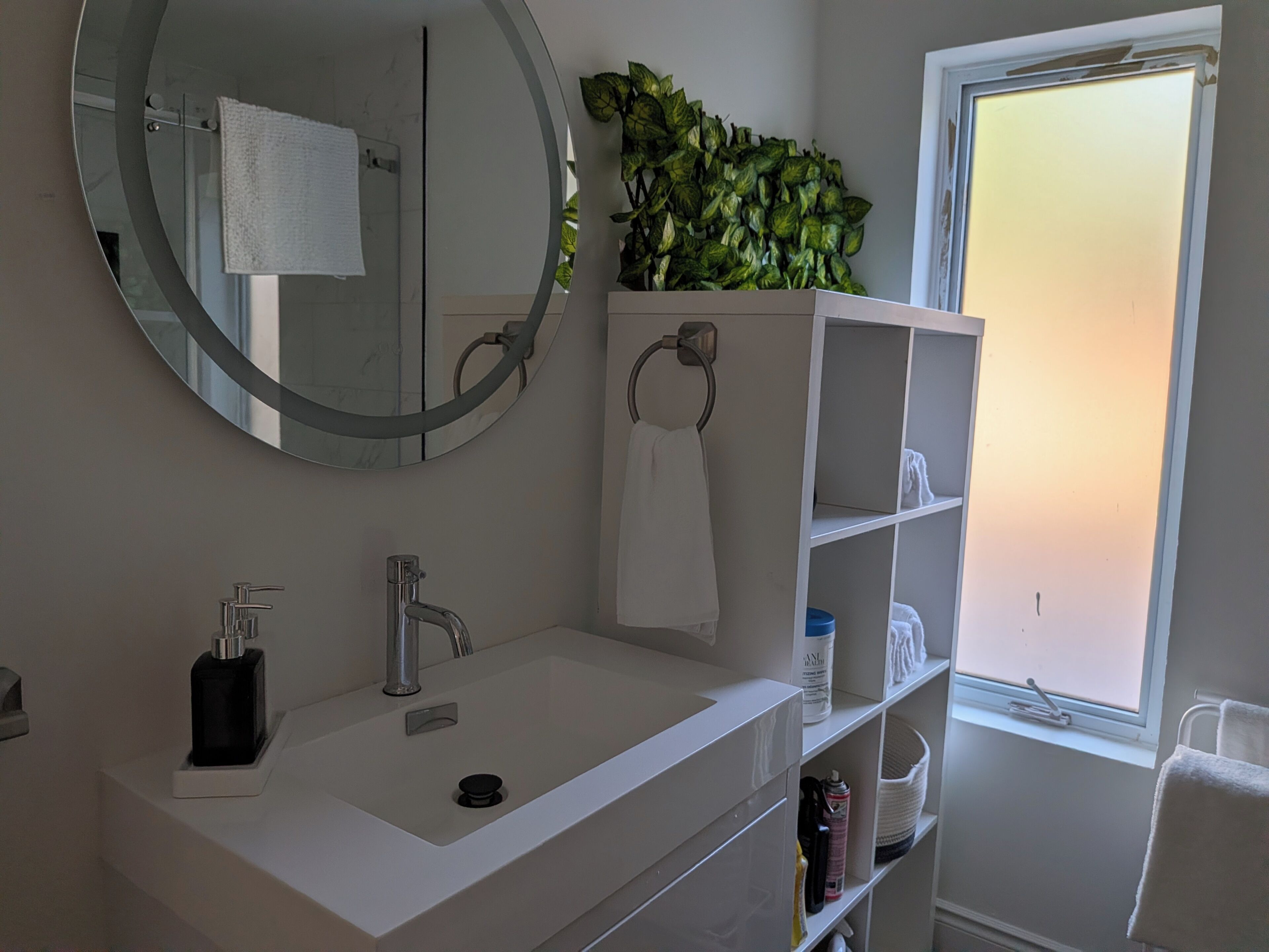 A bathroom with a white sink, a round mirror, a towel rack, a shelf with toiletries, and a window with a view.