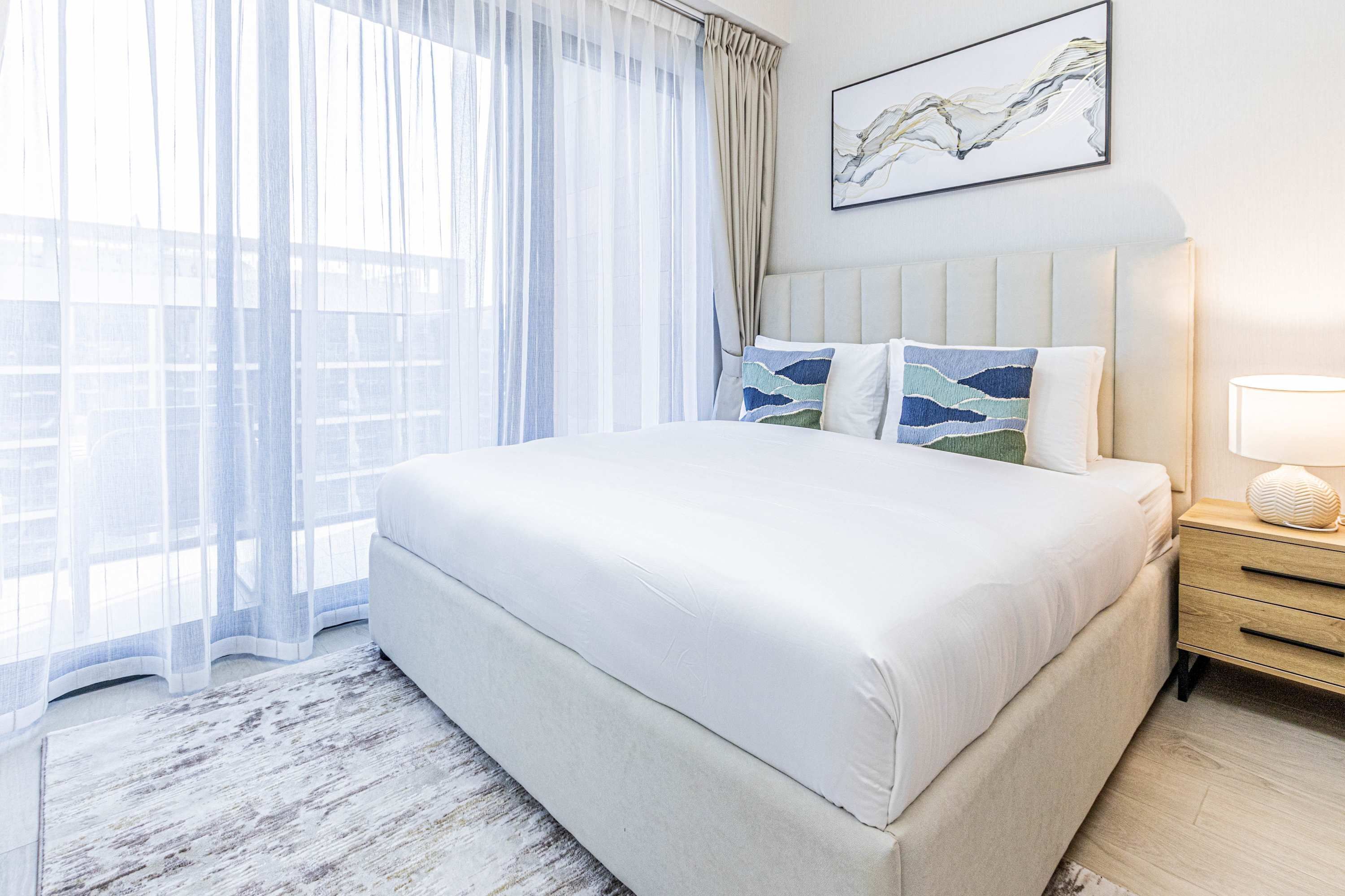 Un lit bien fait, recouvert d’une couvre-lit blanche, avec une tête de lit agrémentée de deux coussins décoratifs, une table de chevet sur laquelle se trouve une lampe, et une fenêtre ornée de rideaux légers.