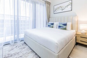 A neatly made bed with a white bedspread, a headboard with two decorative pillows, a bedside table with a lamp, and a window with sheer curtains.