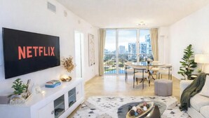 A modern living room with a large TV displaying the Netflix logo, a dining area with a glass table, and a sofa with a view of the cityscape through large windows.