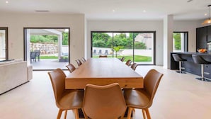 Un vaste espace de salle à manger avec une longue table en bois et des chaises modernes, de grandes portes-fenêtres coulissantes donnant sur un patio extérieur, et une cuisine contemporaine dotée de tabourets de bar.