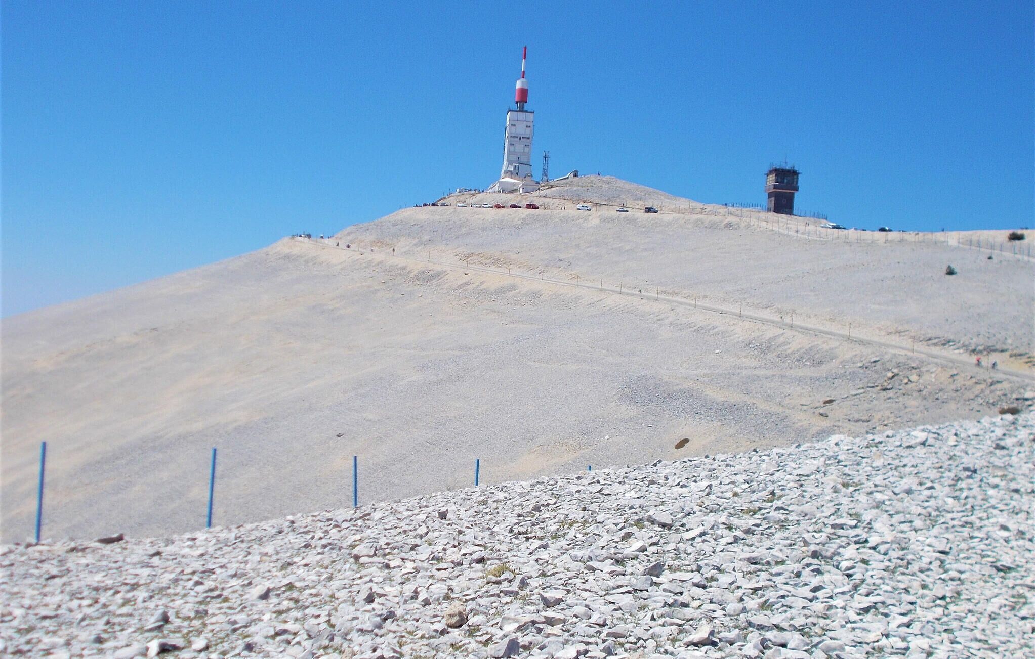 Een kaal heuveltop met een hoge, smalle constructie, herkenbaar aan de rode en witte top, mogelijk een zendmast, en een kleiner gebouw met een rood dak.