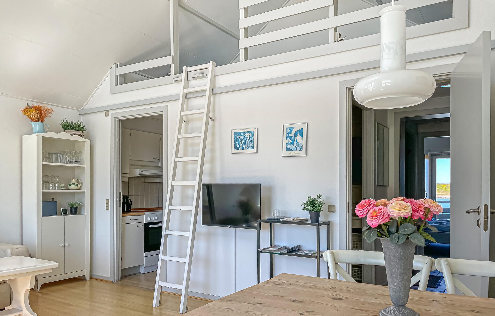 Une cuisine moderne avec des meubles blancs, un four encastré et un téléviseur posé sur un meuble. Une échelle blanche est appuyée contre le mur, et un vase rempli de fleurs se trouve sur la table à manger.