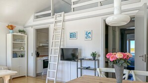 Et moderne køkken med hvide skabe, en indbygget ovn og et fjernsyn på et stativ. En hvid stige står lænet mod væggen, og der er en vase med blomster på spisebordet.