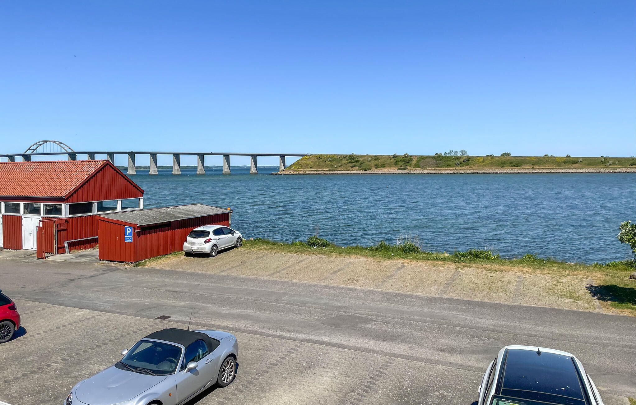 Un estacionamiento con un edificio rojo, un coche blanco y un puente al fondo.