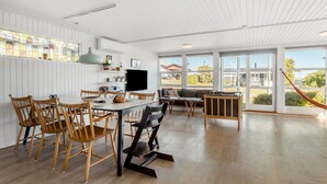 Ein geräumiger Wohnbereich mit Esstisch, Stühlen und einer Couch. Ein Fernseher ist an der Wand befestigt, und große Fenster bieten einen Blick ins Freie.