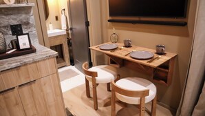 Una cocina compacta con armarios de madera, encimera de mármol, una pequeña mesa de comedor con dos sillas y un televisor montado en la pared.
