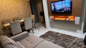 A modern living room with a flat-screen TV mounted on a wooden wall, a grey sofa, and a dining area with a table and chairs.