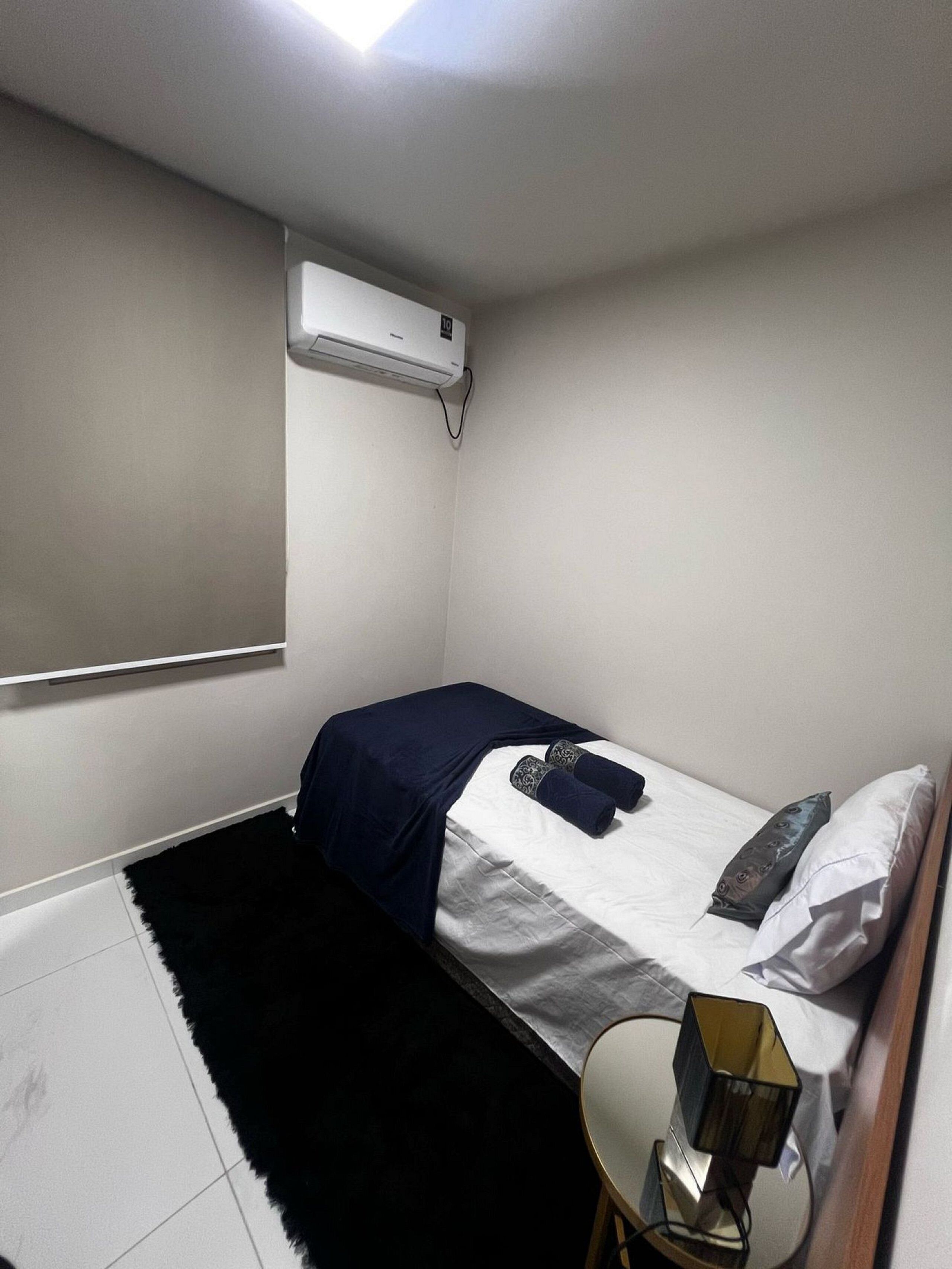 Quarto pequeno e bem iluminado, com uma cama de solteiro, mesa de cabeceira e ar condicionado fixado na parede.