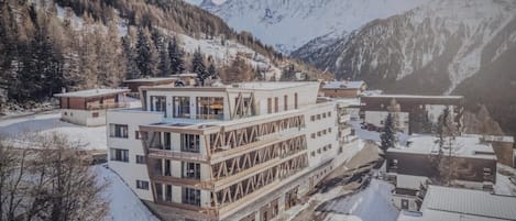 Un hotel moderno con una estructura de varios pisos, situado en una zona montañosa nevada y con vistas a picos circundantes.
