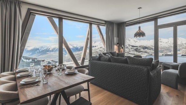 Un comedor moderno con una mesa de madera preparada para cuatro personas, un sofá y vistas a montañas nevadas.