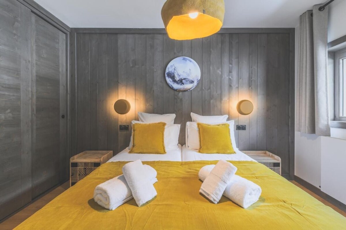 A hotel room with a wooden panel wall, a bed with yellow bedding and pillows, two bedside tables with lamps, and a round wall decoration.