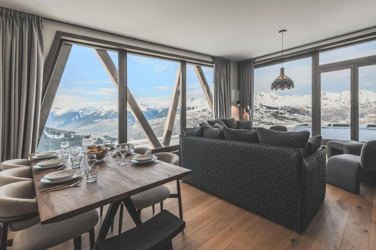 A modern dining area with a wooden table set for four, a sofa, and a view of snow-covered mountains.