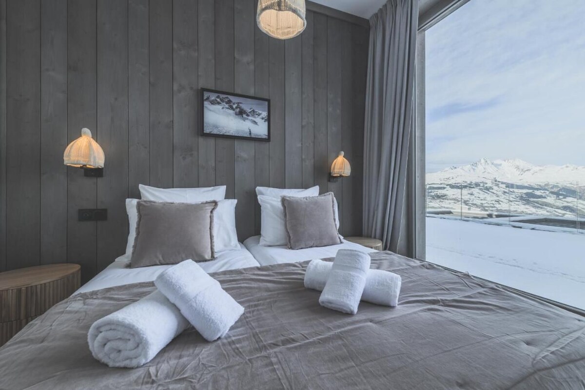 A modern hotel room with a large bed, two wall-mounted lamps, a framed picture, and a view of snow-covered mountains.