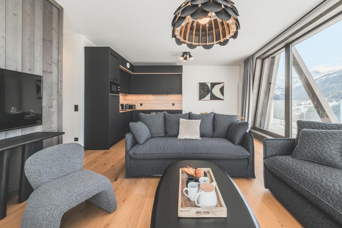 Ein modernes Wohnzimmer mit einem grauen Sofa, einem Holztisch mit einer Tablett voller Gegenstände und einem großen Fenster mit Blick auf Berge.