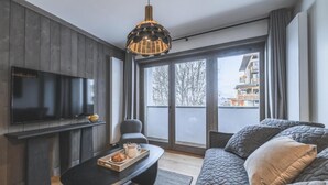 Una sala de estar moderna con un televisor de pantalla plana, una mesa de centro negra con una bandeja de comida, un sofá gris y un ventanal con vistas a edificios.