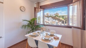 Un coin repas avec une table dressée pour deux personnes, une horloge au mur et une vue sur une ville perchée sur une colline, visible à travers une fenêtre.