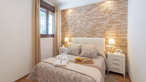 Une chambre à coucher bien aménagée, avec un mur en pierre, un lit recouvert d’une couverture beige, deux tables de chevet avec des lampes et un plateau de fruits posé sur le lit.