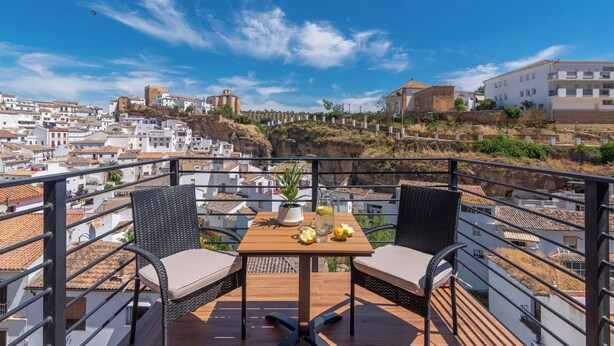 Un balcon avec deux fauteuils en osier, une table en bois sur laquelle est posée une carafe de limonade, et une vue sur une ville côtière aux bâtiments blancs et à la forteresse historique.