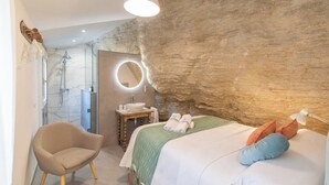 A bedroom with a bed, a chair, a vanity with a mirror, and a bathroom with a shower.
