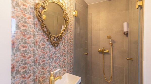 A bathroom with a floral-patterned wall, a gold-framed mirror, a sink, and a shower with a gold faucet and hose.