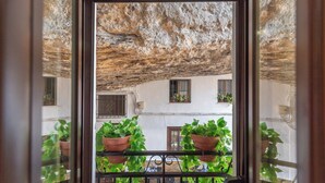 A balcony with potted plants, a view of a building with white walls and multiple windows, and a stone wall above. - Bajo la Roca Setenil de las Bodegas by Ruralidays (Cádiz)