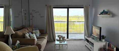 A living room with a sofa, a coffee table, a TV, and a balcony with a view of a green landscape.