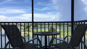 Ein Balkon mit zwei Korbstühlen und einem kleinen runden Tisch, mit Blick auf einen Golfplatz und ein Gewässer.