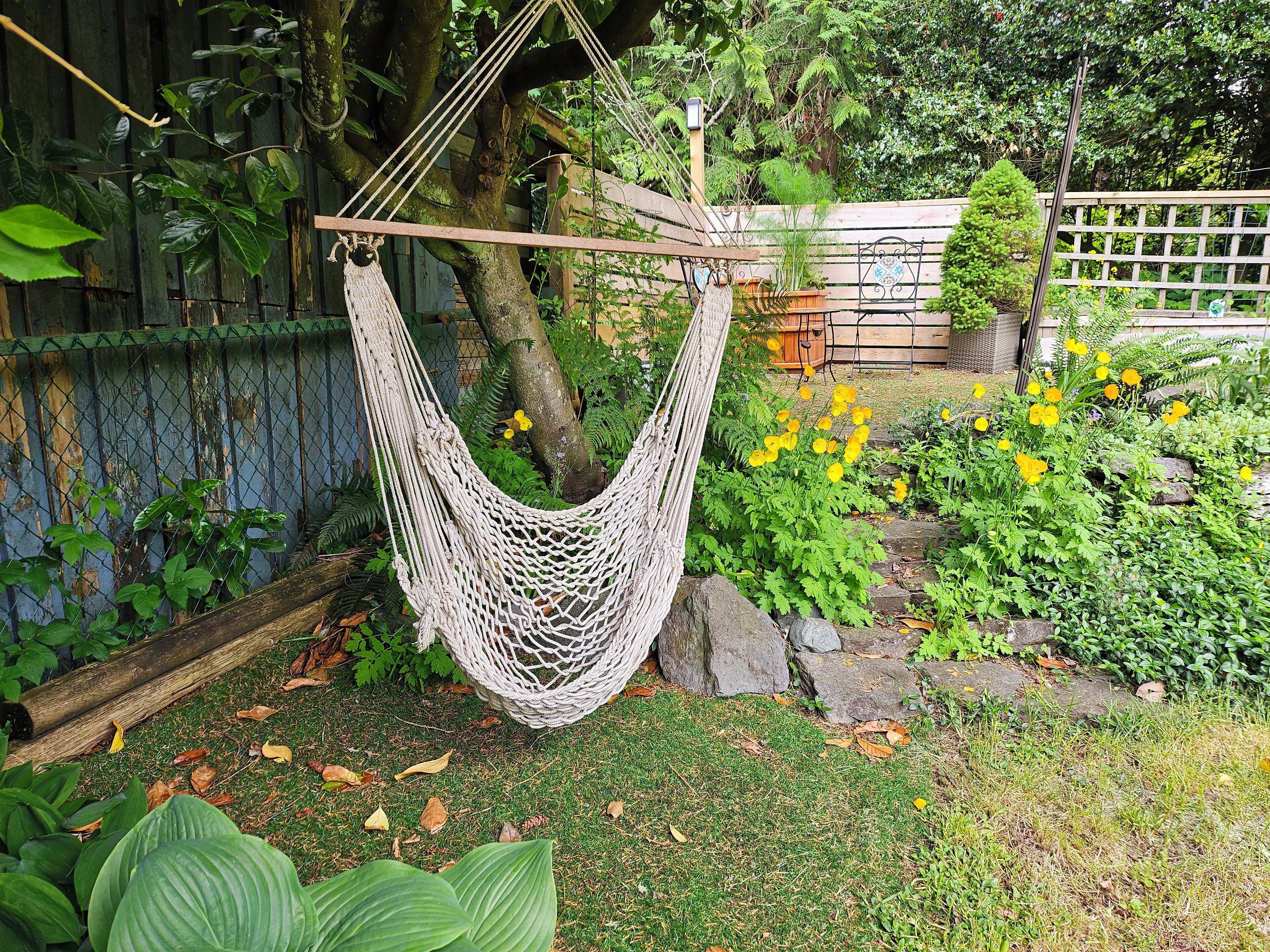 Un hamac suspendu entre des arbres dans un jardin, avec une clôture en bois, un chemin de pierres et diverses plantes et fleurs.