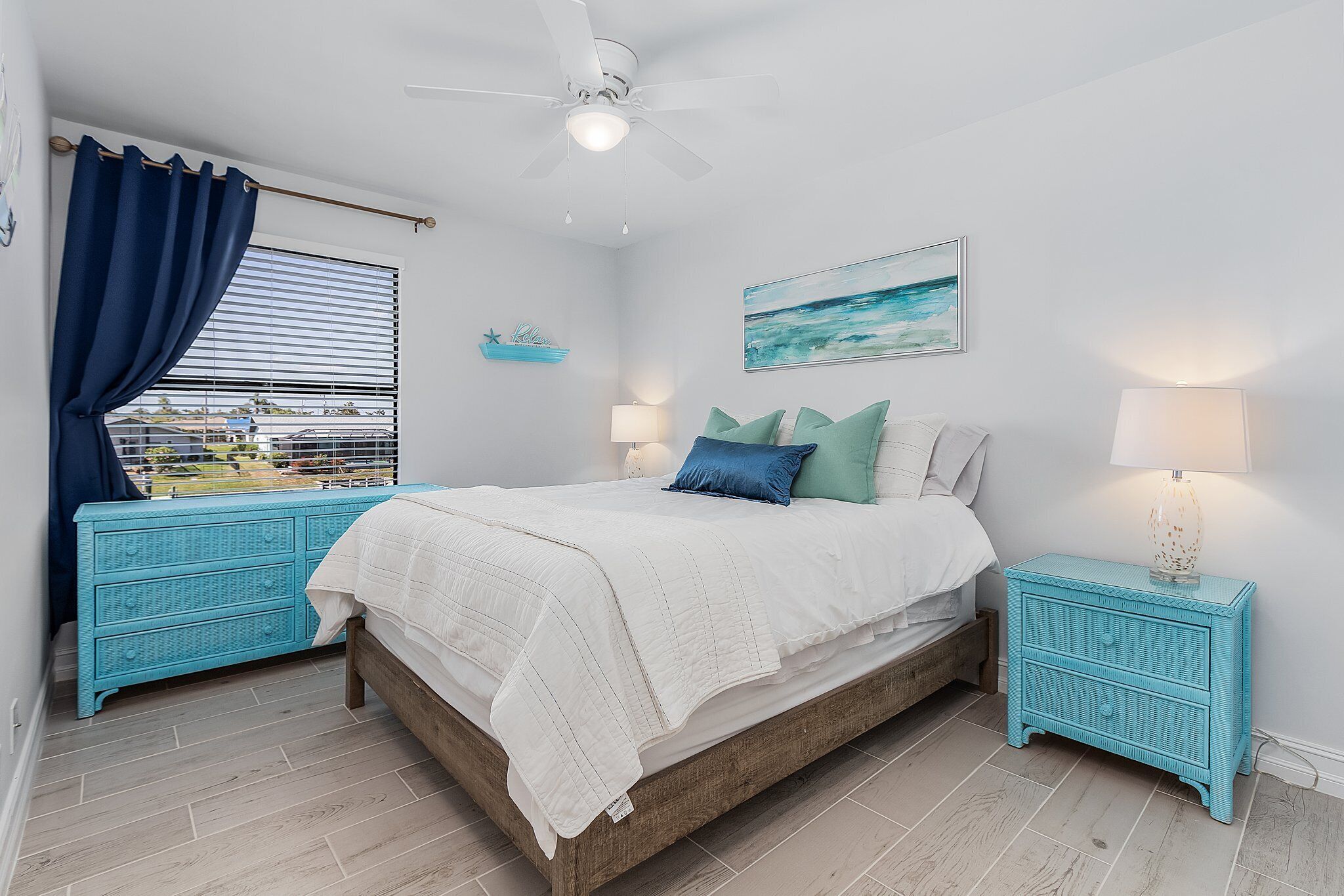 Een slaapkamer met een bed, twee nachtkastjes, een plafondventilator, een raam met gordijnen, een wandplank en een schilderij.