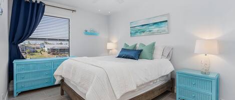 Een slaapkamer met een bed, twee nachtkastjes, een plafondventilator, een raam met gordijnen, een wandplank en een schilderij.