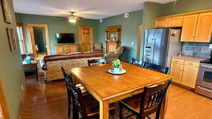 Un comedor con una mesa y sillas de madera, un televisor al fondo y armarios de madera en la cocina.