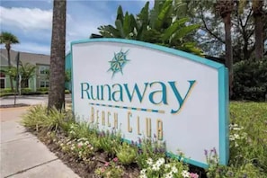 Ein Schild für den Runaway Beach Club mit einem Stern-Emblem, umgeben von Grünpflanzen und einem Bürgersteig.