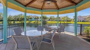 A covered outdoor patio with round tables and chairs, a ceiling fan, and a view of a residential area with houses and a body of water. - Sunshine Paradise in lovely Kissimmee with WiFi (Kissimmee)