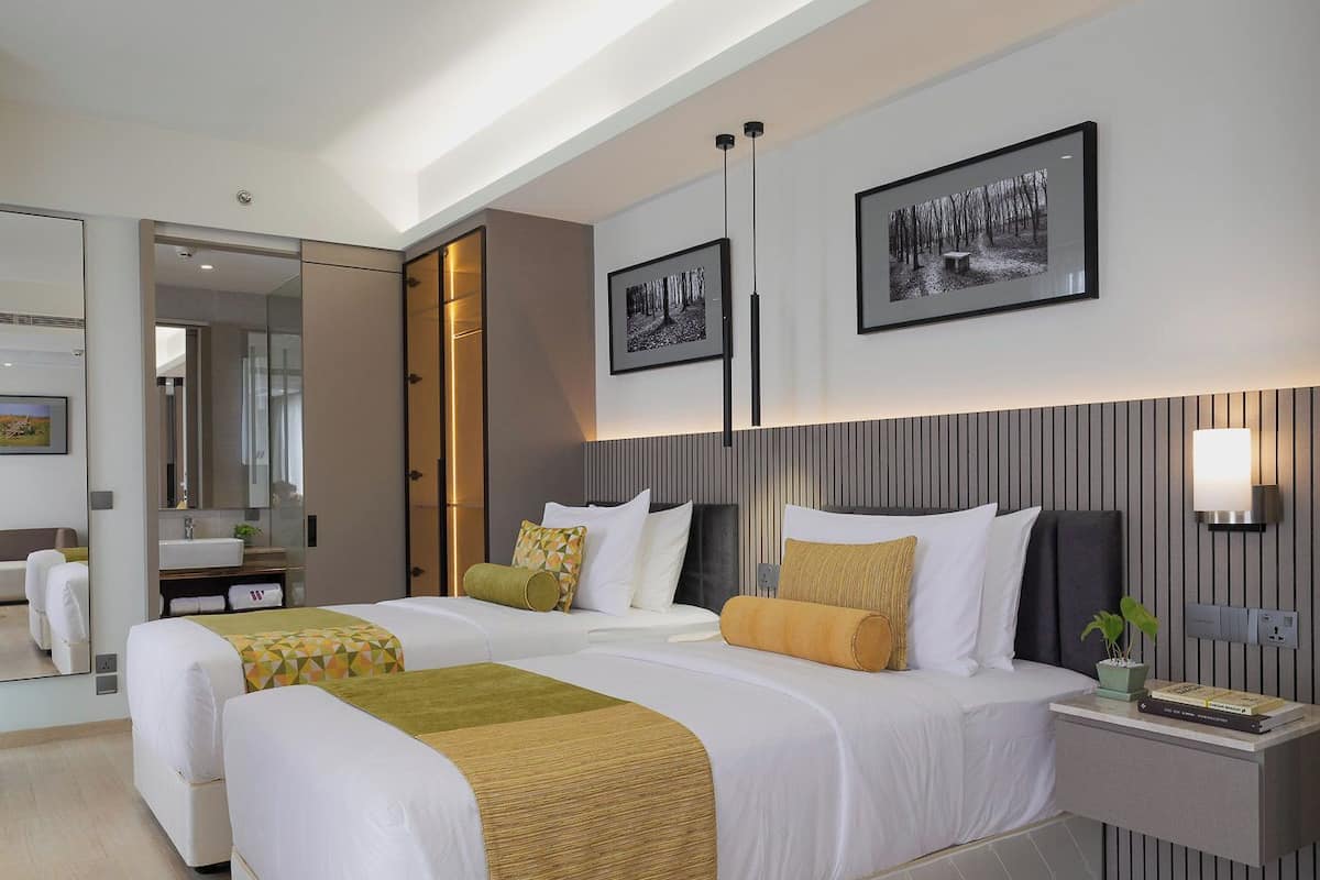 Una habitación de hotel moderna con dos camas, un espejo grande, una mesita de noche con lámpara y cuadros enmarcados en la pared.