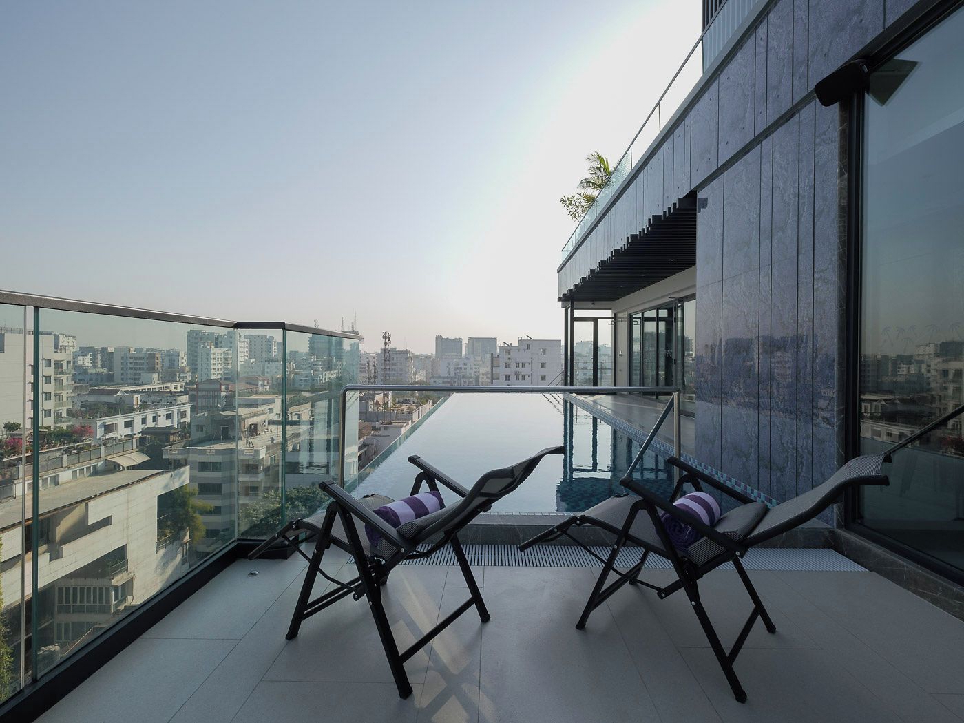 Un balcón moderno con barandillas de vidrio, dos sillones de descanso y vistas a un paisaje urbano.