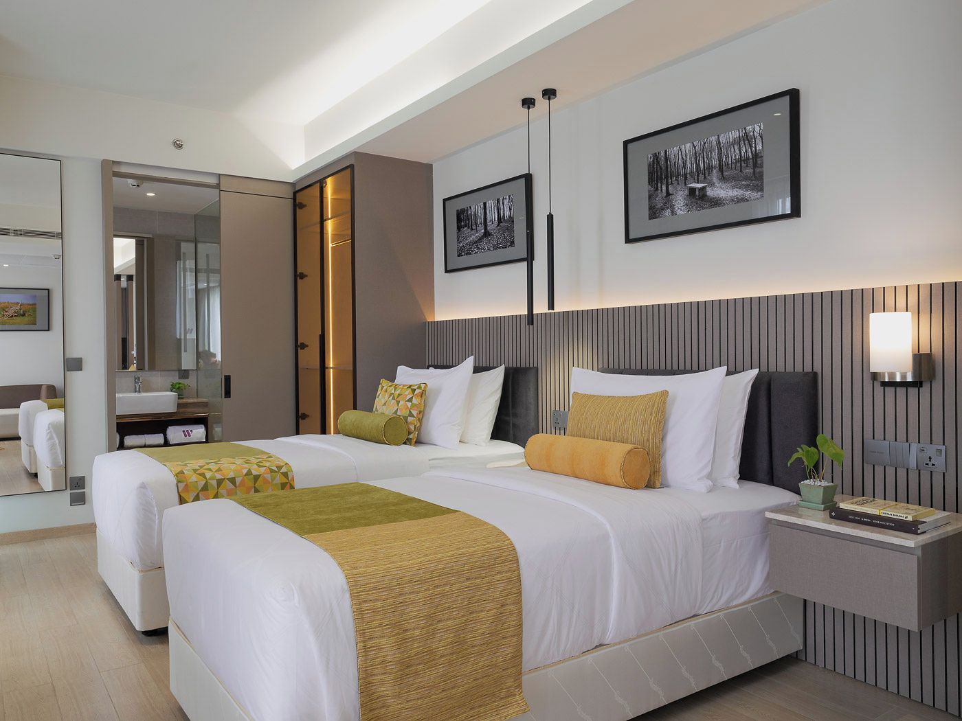 A modern hotel room with two beds, a large mirror, a bedside table with a lamp, and framed pictures on the wall.