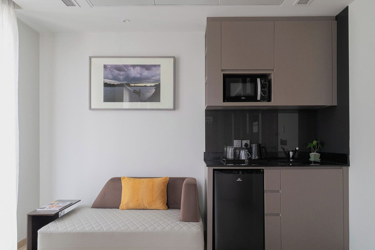A compact kitchenette with a microwave, coffee maker, and small refrigerator. A sofa with a yellow pillow is positioned in front of the kitchenette.