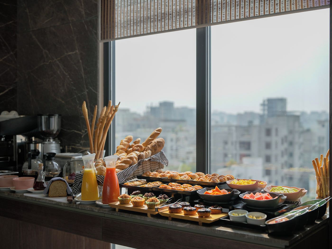 A breakfast buffet with various pastries, fruits, and beverages, set against a city view.