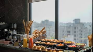 A breakfast buffet with various pastries, fruits, and beverages, set against a city view.