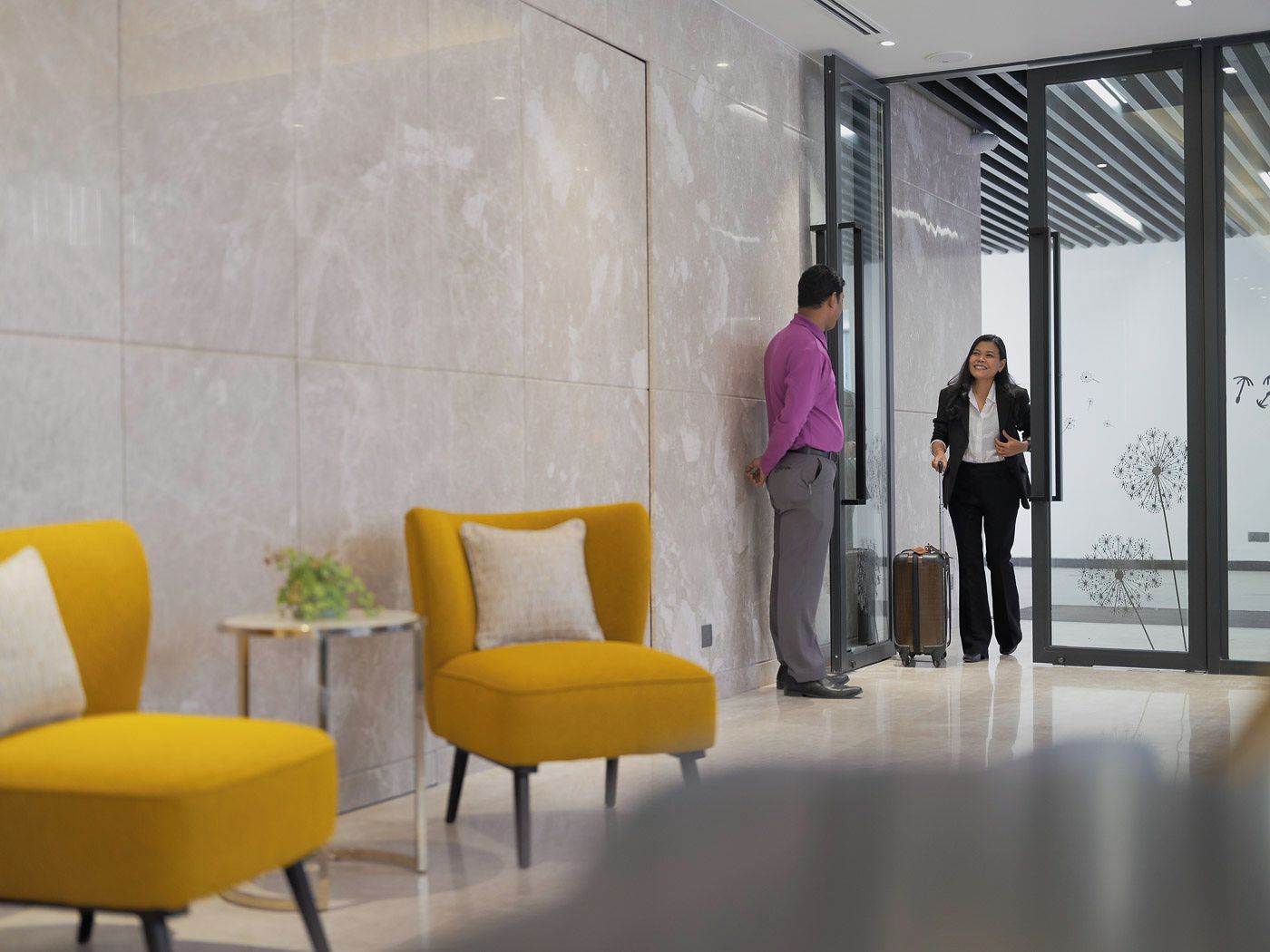 A modern lobby with marble walls, a glass door entrance, and yellow armchairs.