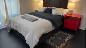 Uma cama bem arrumada com uma toalha cinzenta, um criado-mudo vermelho com uma luminária e um tapete estampado.