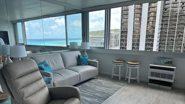A modern living room with a large window overlooking the ocean, a sofa, two side chairs, a small rug, and a wall-mounted air conditioner. - Cozy studio in Waikiki  across the street from Waikiki Beach (Honolulu)